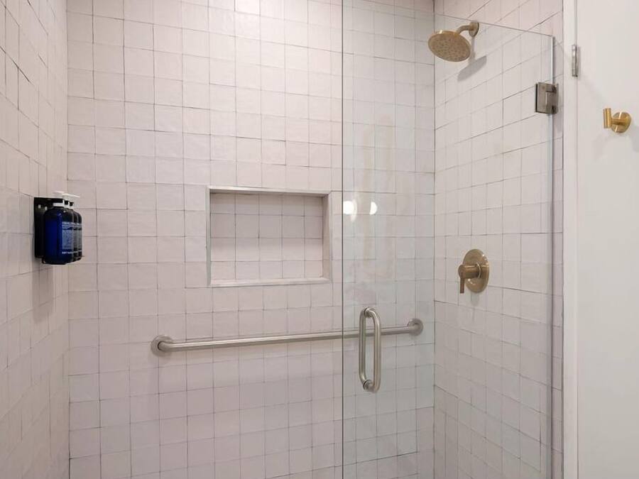 A shower area with white tiled walls, a glass door, a handheld showerhead, a grab bar, and a soap dispenser.