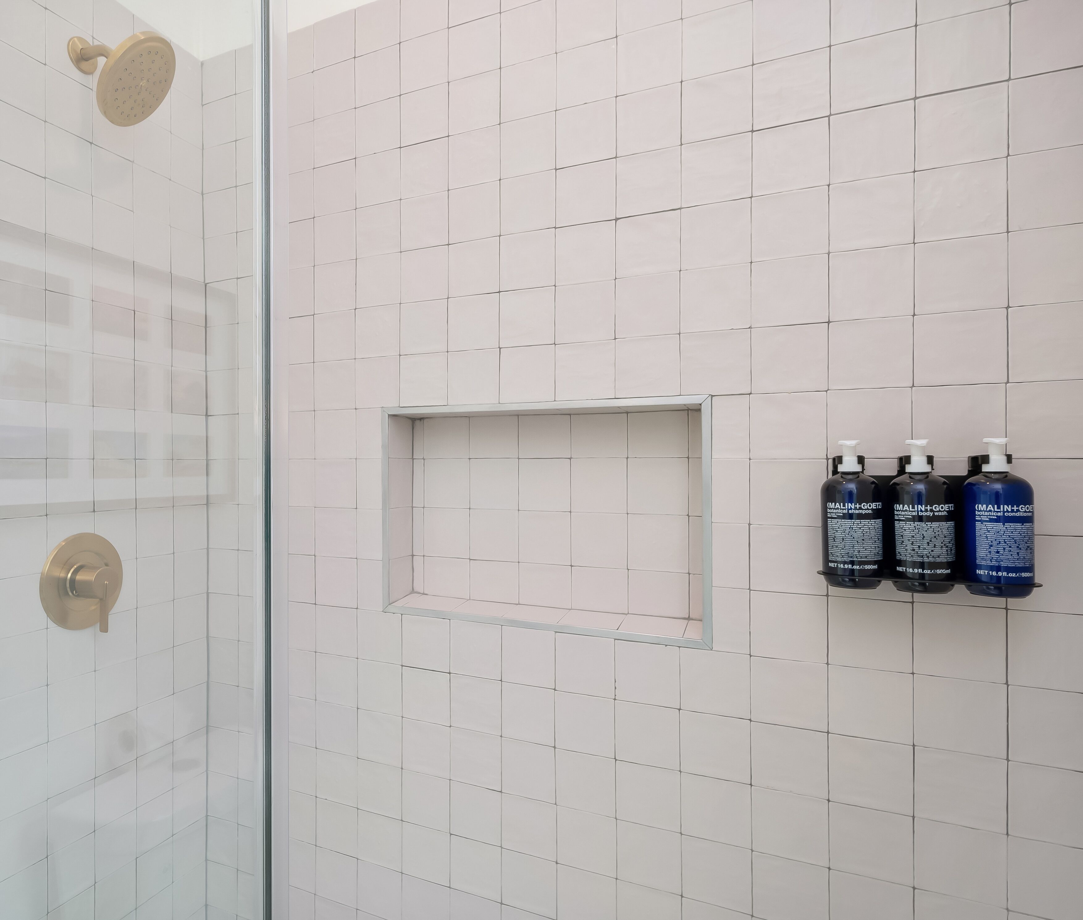 A bathroom with white tiles, a shower with a gold handle, and three bottles of Malini+Goeti body wash on the wall.
