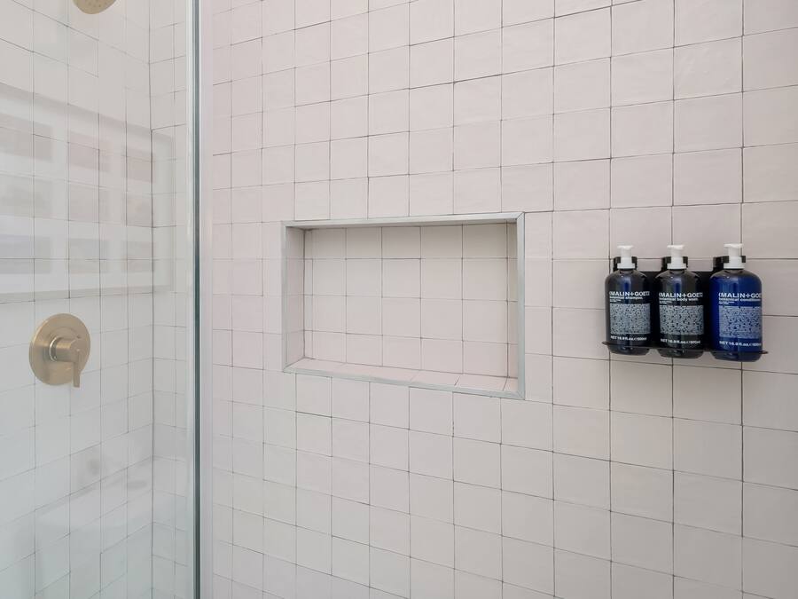 A bathroom with white tiles, a shower with a gold handle, and three bottles of Malini+Goeti body wash on the wall.