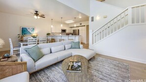 A spacious living room with a large L-shaped sofa, a coffee table, and a dining area with a kitchen in the background. - Charming and immaculate, fully furnished 3-bedroom 3 bath townhouse in Ko'Olina! (Kapolei)