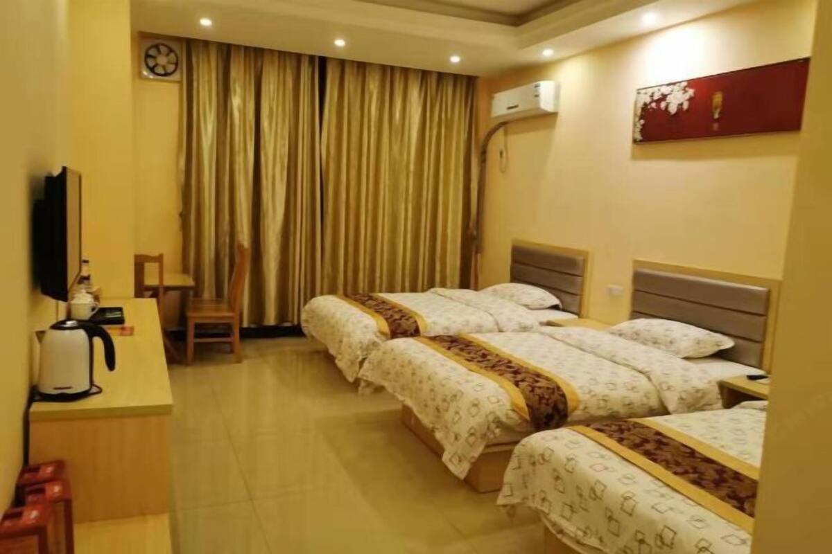A hotel room with three beds, a desk with a kettle, a chair, a clock, and an air conditioning unit.