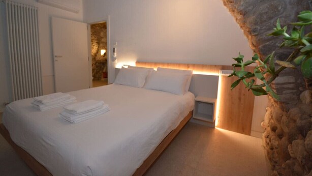 A bedroom with a bed, white linens, a wooden headboard, a stone wall, and a small plant. - Residence Corte Bellebarbe - Il Volto (rivoli veronese)