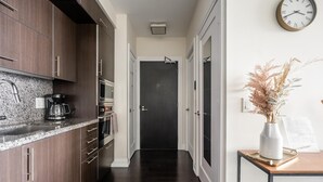 Een moderne keuken met donkerhouten kasten, een granieten aanrechtblad en roestvrijstalen apparatuur. Er is een gang die naar een donkere deur leidt, en een klein tafeltje met een vaas gedroogde bloemen aan de rechterkant.