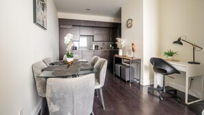 Een moderne keuken met donkere kastjes, een eethoek met een glazen tafel en grijze stoelen, en een klein bureau met een lamp.