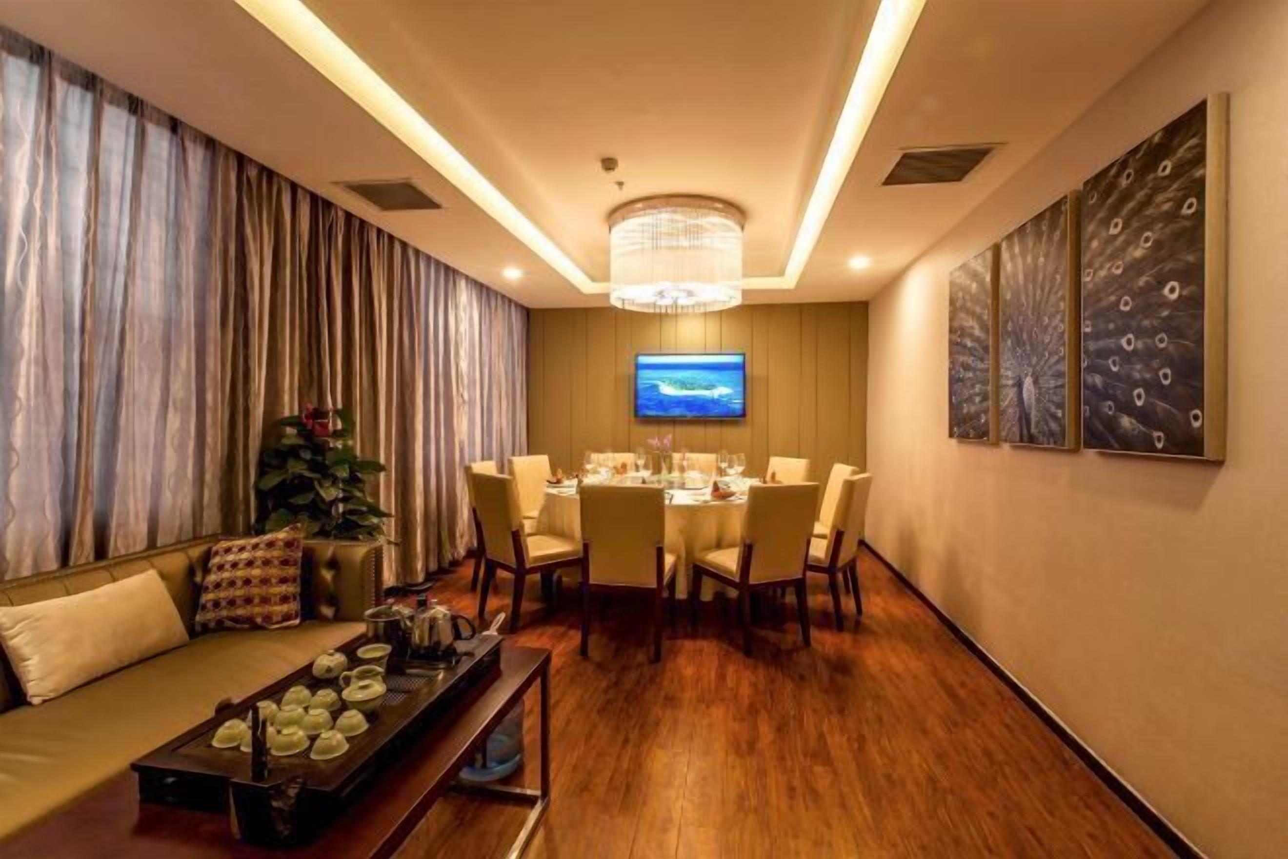A modern dining area with a round table set for six, a sofa with a tray of teapots, and a television on the wall.