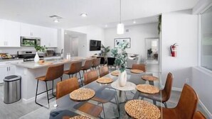 Une cuisine moderne avec des armoires blanches, une cuisinière en acier inoxydable et un coin repas comprenant une table en verre et des sets de table en fibres tressées.