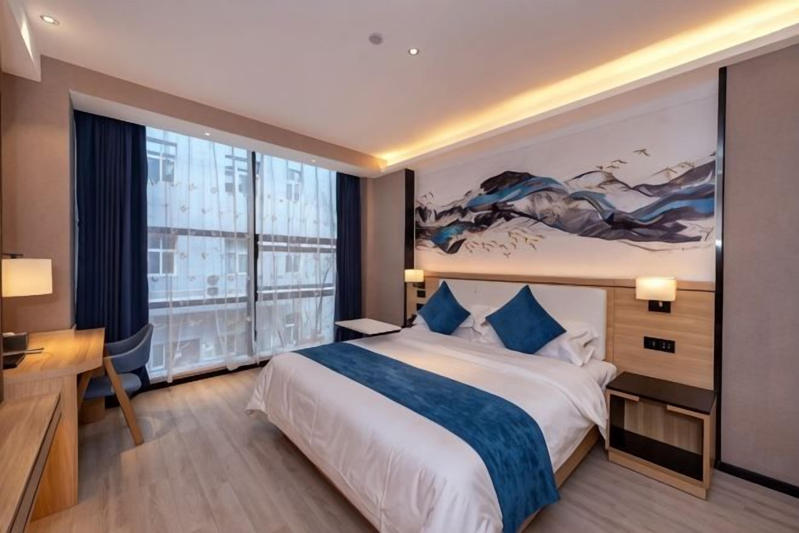 A modern hotel room with a large bed, bedside tables, a desk, and a view through a large window.