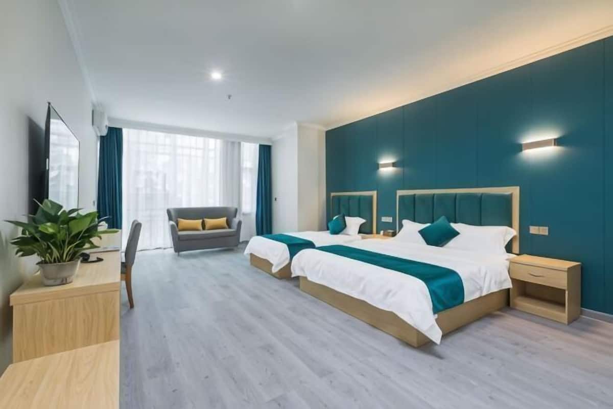 A modern hotel room with two beds, a desk, a chair, a potted plant, and a view of the city.
