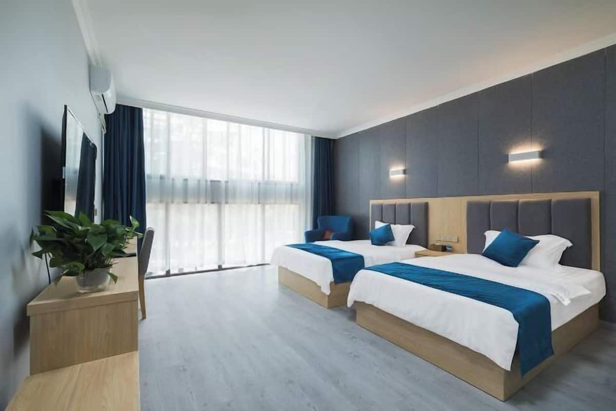 A hotel room with two beds, a desk, a plant, and a large window.