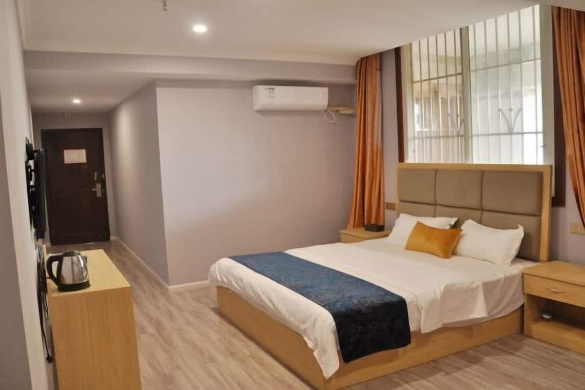 A modern hotel room with a large bed, a bedside table, a nightstand, an air conditioning unit, and a window with curtains.
