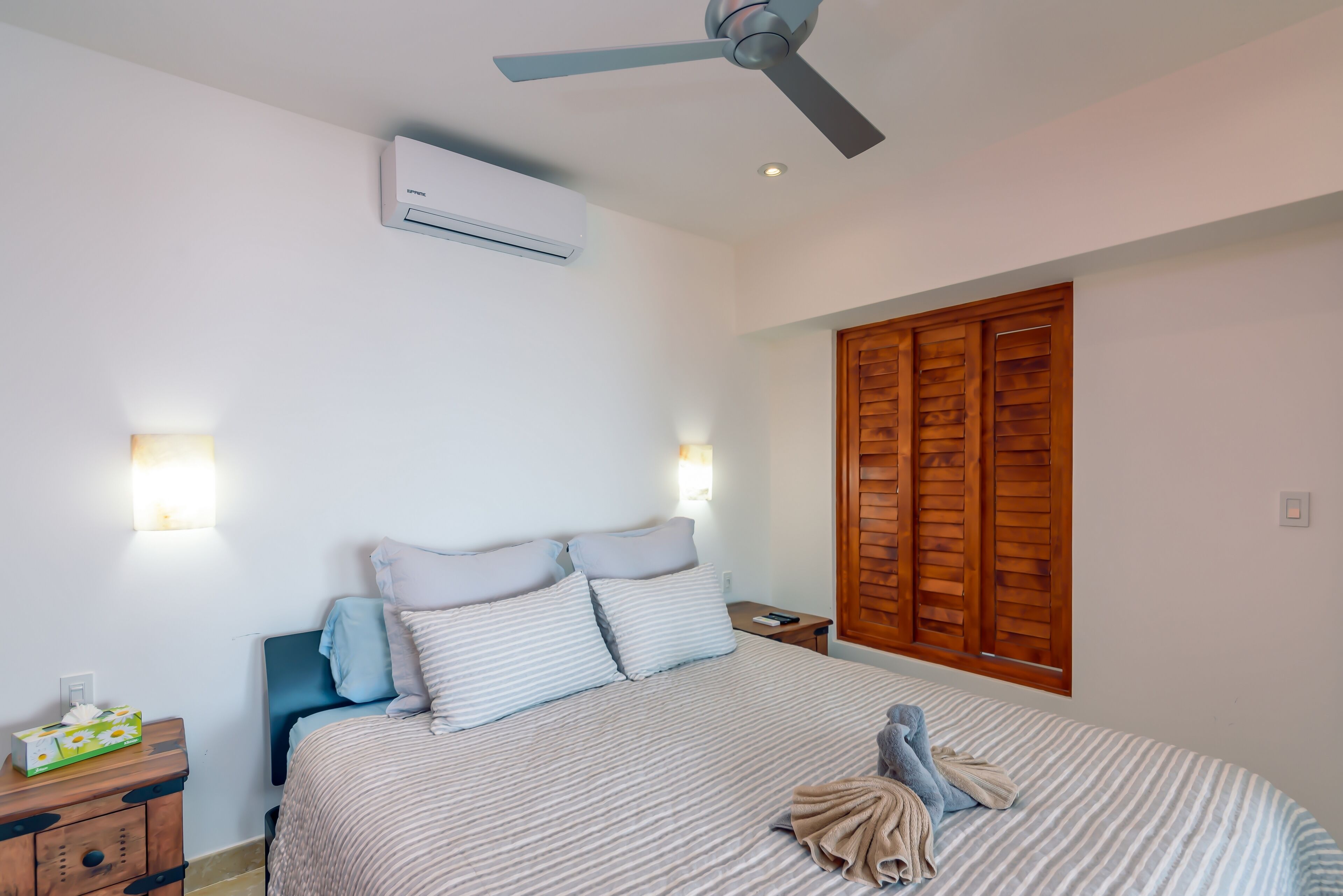 Une chambre à coucher comprenant un lit, une table de chevet, une commode, un climatiseur mural, un ventilateur de plafond et des volets en bois.
