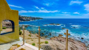A coastal view with a rocky shoreline, a clear blue sky, and a yellow building with an arched window. - Oceanfront, Gound floor, Steps to the Beach!  (Cabo San Lucas)