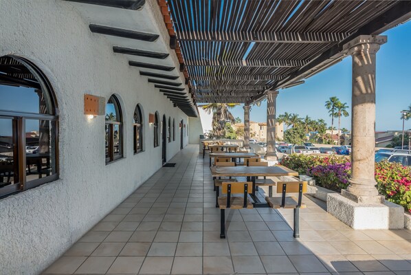 Un patio al aire libre con mesas y sillas de madera, una pérgola y vistas a coches aparcados y palmeras.