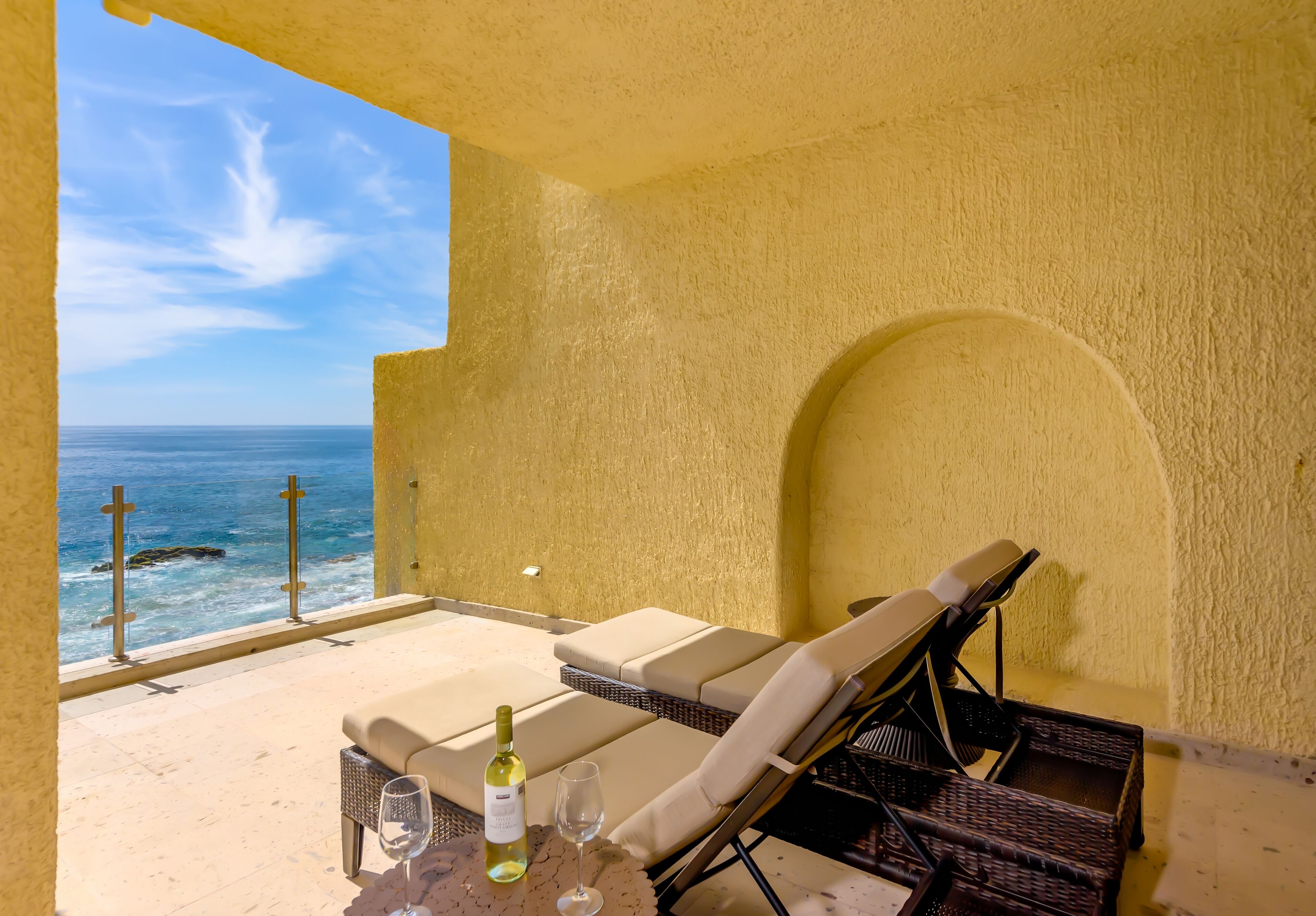 A balcony with a view of the ocean, a wicker lounge chair, a bottle of wine, and a glass on a table.