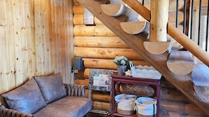 L’intérieur d’une cabane en rondins, avec un escalier en bois, un canapé, une petite table agrémentée d’objets décoratifs et une cheminée.