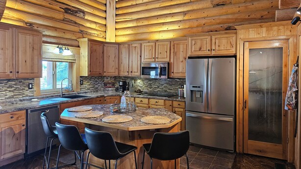 Une cuisine spacieuse avec des meubles en bois, un grand îlot central doté de tabourets, des appareils électroménagers en acier inoxydable et une vue sur l’extérieur grâce à une fenêtre.