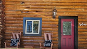 Une cabane en rondins avec une porte rouge, une pancarte indiquant « Bienvenue » et deux chaises portant l’inscription « véranda ».