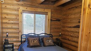 Une chambre à coucher située dans une cabane en rondins, avec un lit, deux tables de chevet, une fenêtre avec des stores et un luminaire au plafond.