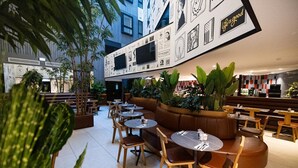 A modern indoor dining area with round tables and benches, surrounded by potted plants and featuring a wall with various black and white images. - So City Hotel - Adults Only (St. Julian's)