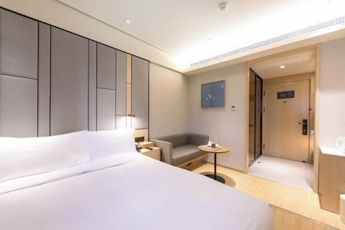 Una habitación de hotel moderna con una cama grande, un pequeño sofá, una mesa redonda y un baño con puerta de madera.