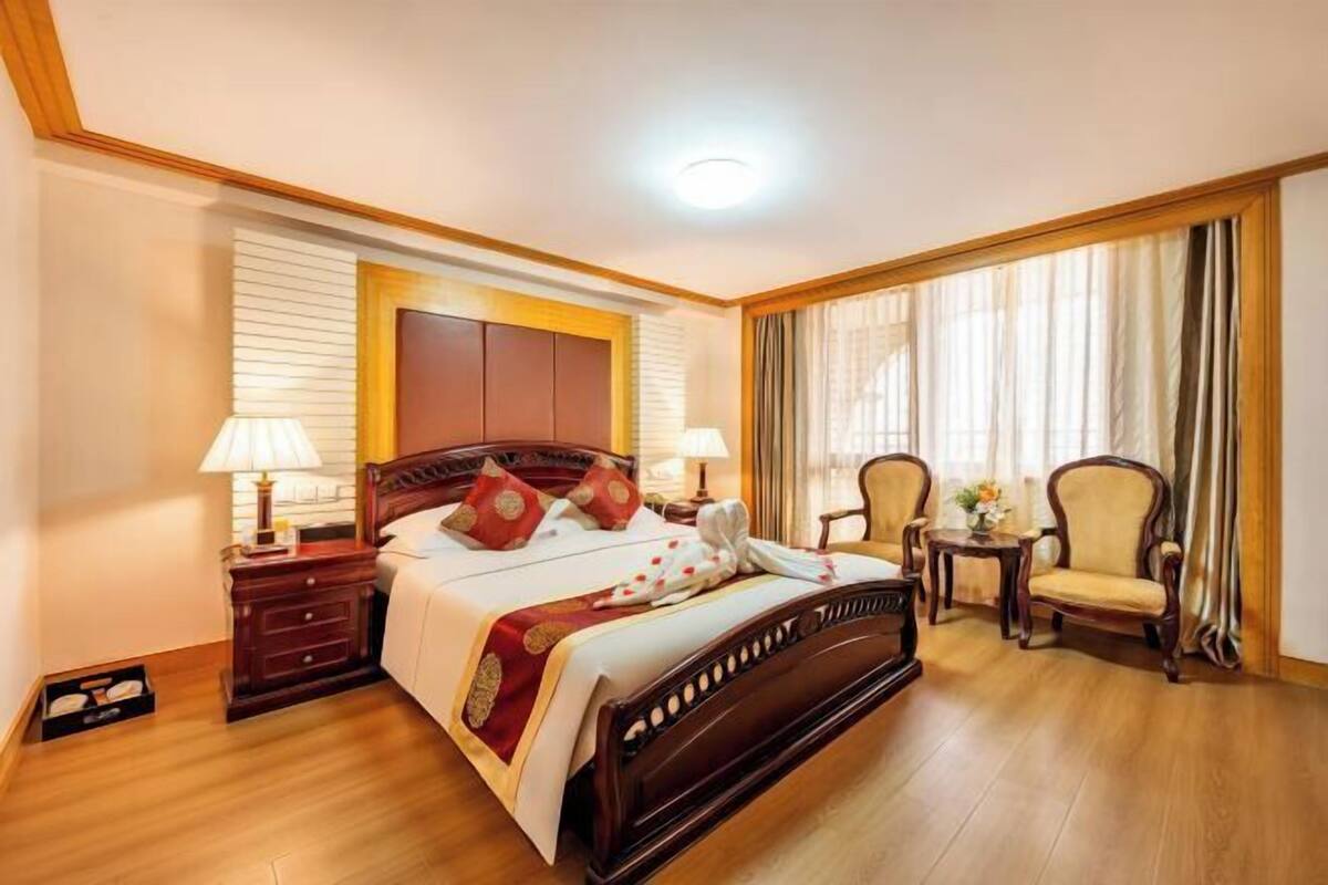 A hotel room with a wooden bed, two bedside tables, a chair, a small table, and a window with curtains.