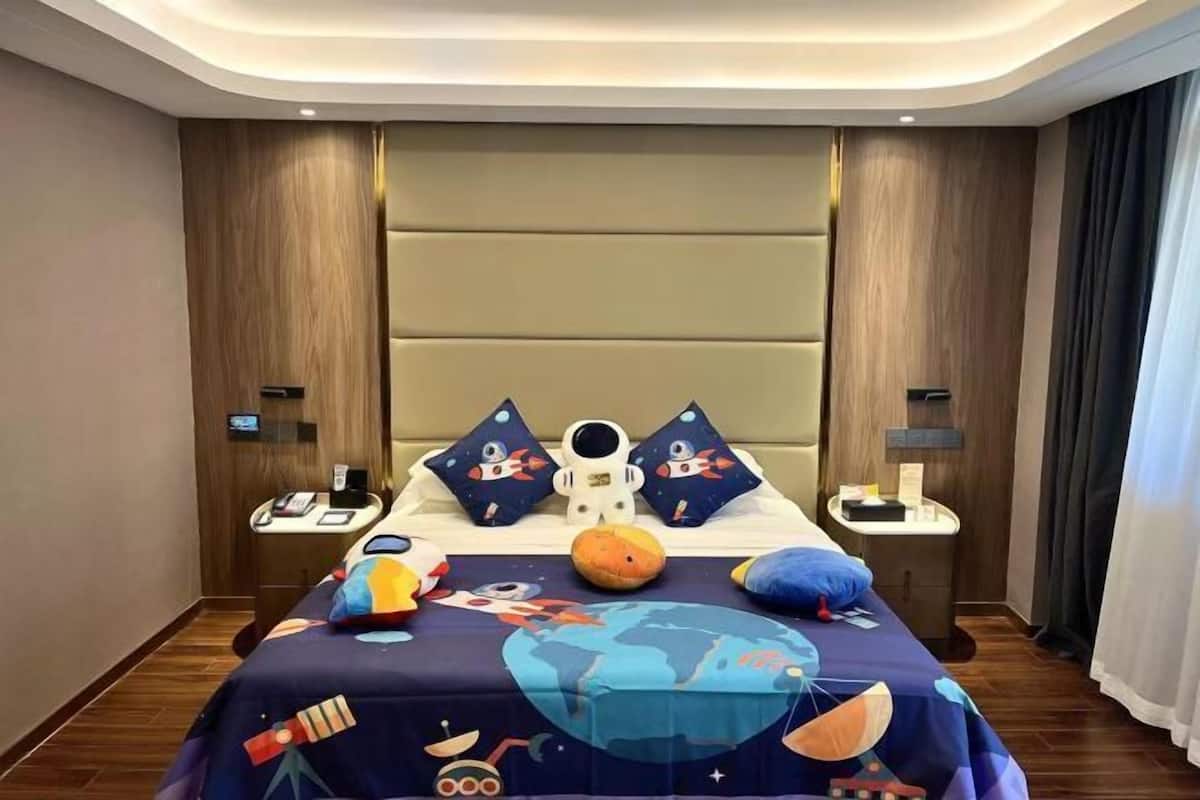 A hotel room with a bed featuring a space-themed bedspread, two nightstands with lamps, and a large window with curtains.