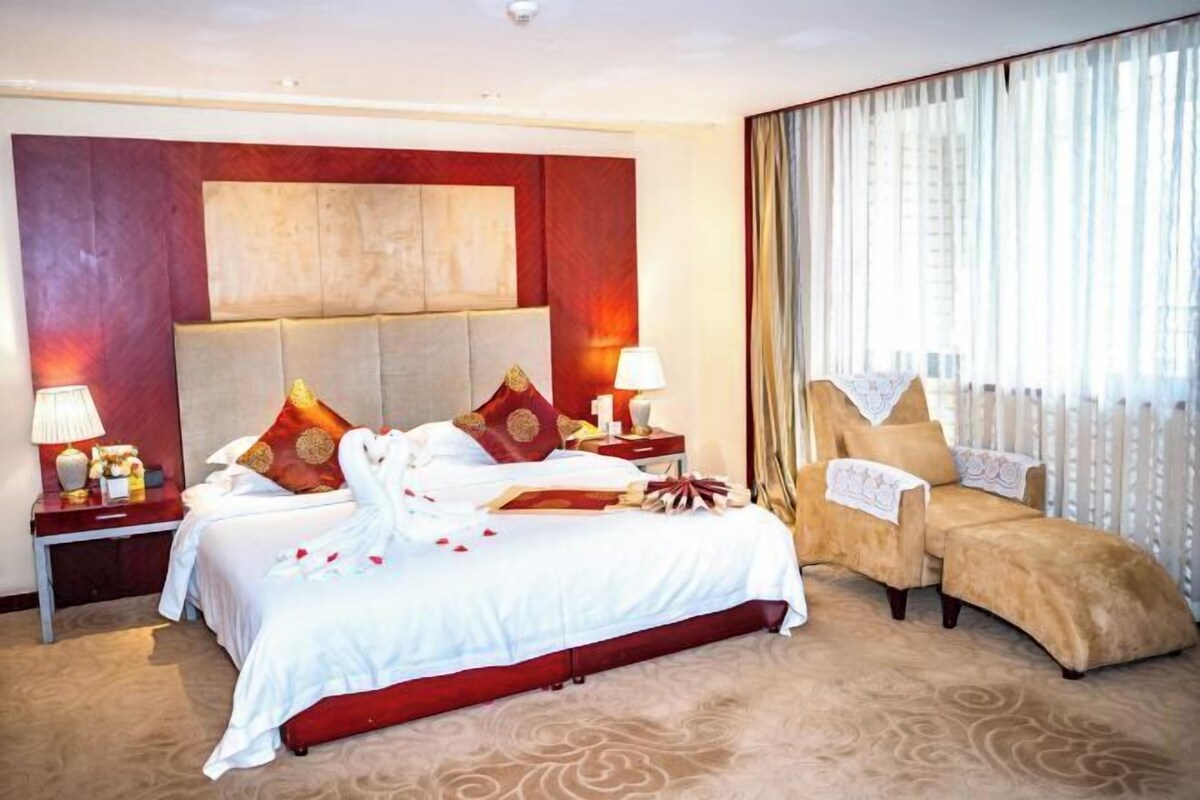 Ein Hotelzimmer mit einem großen Bett, zwei Nachttischen, einem Sessel und einem großen Fenster mit Vorhängen.