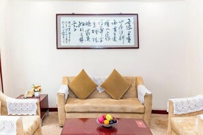 Ein Zimmer mit einem beigefarbenen Sofa, zwei passenden Ohrensesseln, einem kleinen Tisch mit Obstschale und einem gerahmten Kalligraphie-Kunstwerk an der Wand.