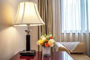 Ein Hotelzimmer mit einer Lampe auf einem Holztisch, einem Blumenstrauß und Blick durch Vorhänge nach draußen.