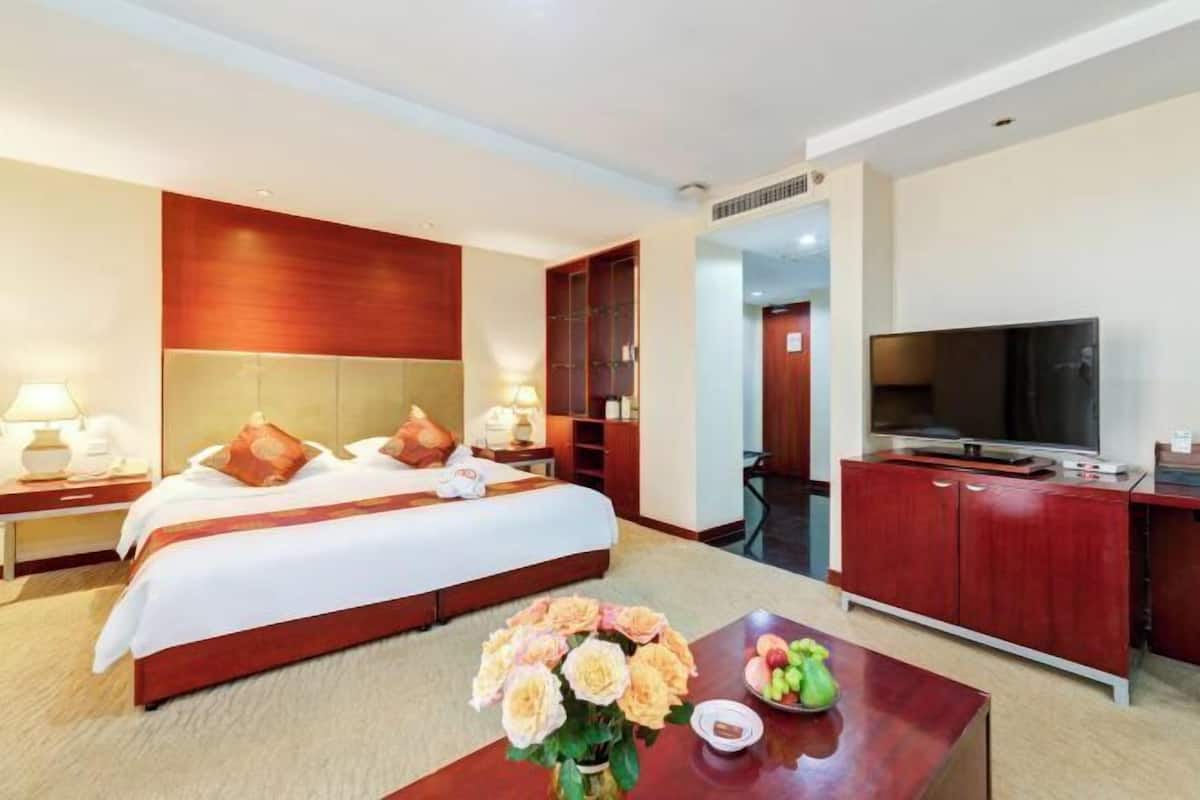 A hotel room with a large bed, a TV, a desk, and a coffee table with flowers and fruit.