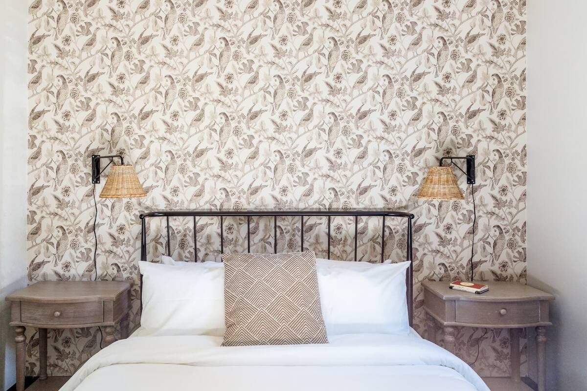 A bedroom with a floral wallpaper, a metal bed frame, two bedside tables with lamps, and a bedside drawer.