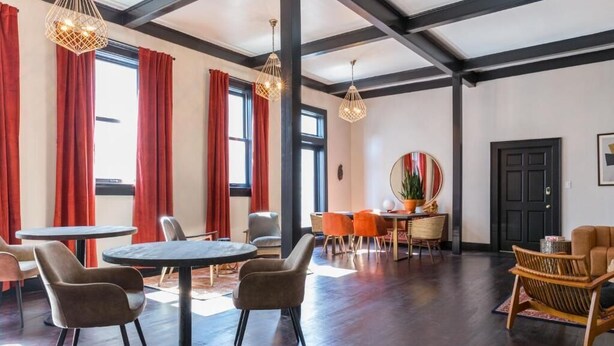 A spacious room with a high ceiling, large windows with red curtains, a round table with chairs, a sofa, and a dining area with orange chairs. - Inn at Gough (San Francisco)