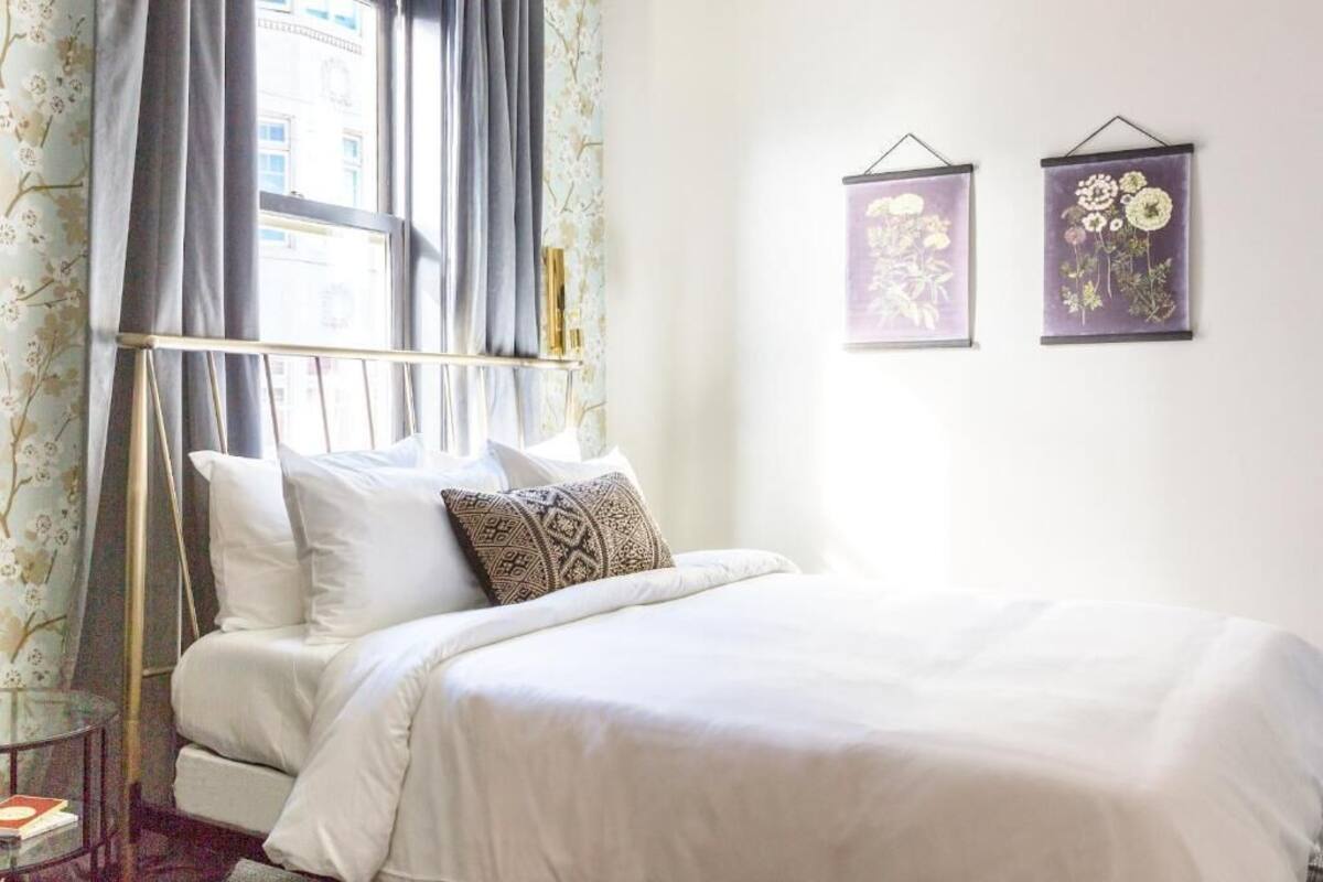 A neatly made bed with white linens and a patterned pillow, a bedside table with a book, and two wall-mounted floral artwork pieces.
