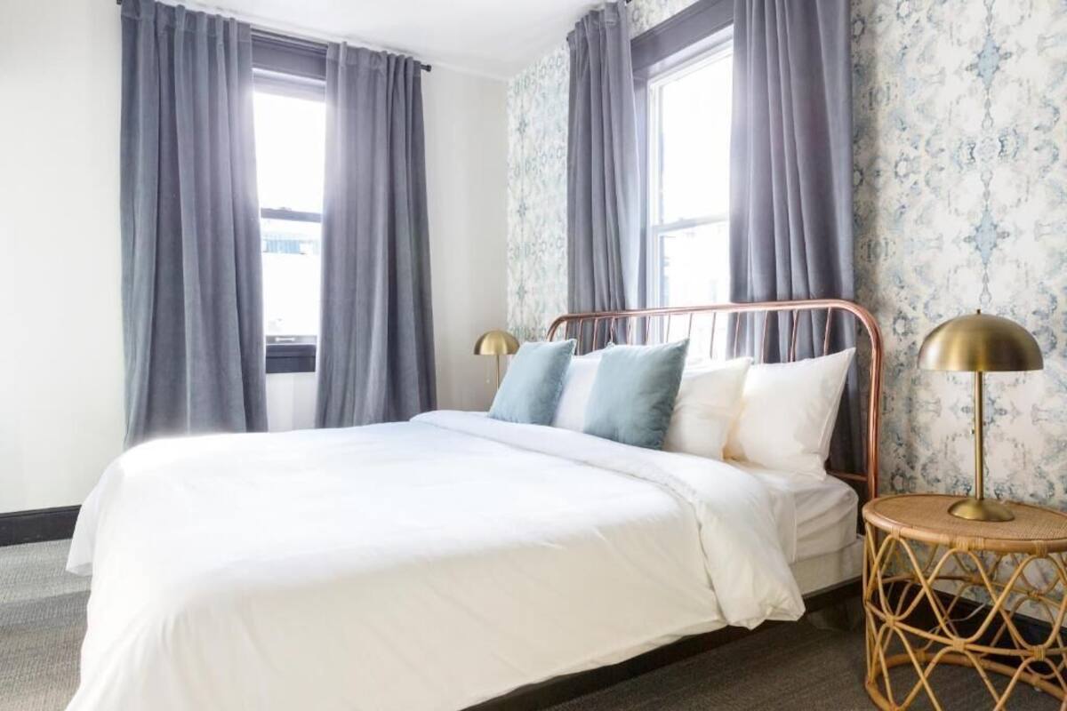 A neatly made bed with a white duvet, grey curtains, and a brass lamp on a round wicker side table.