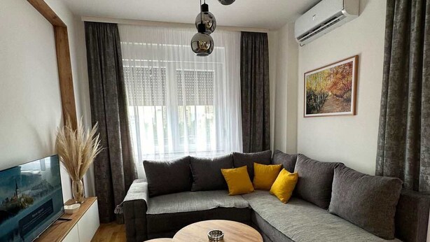 Un salon moderne avec un canapé d’angle, deux petites tables rondes, un téléviseur à écran plat et une grande fenêtre ornée de rideaux légers.