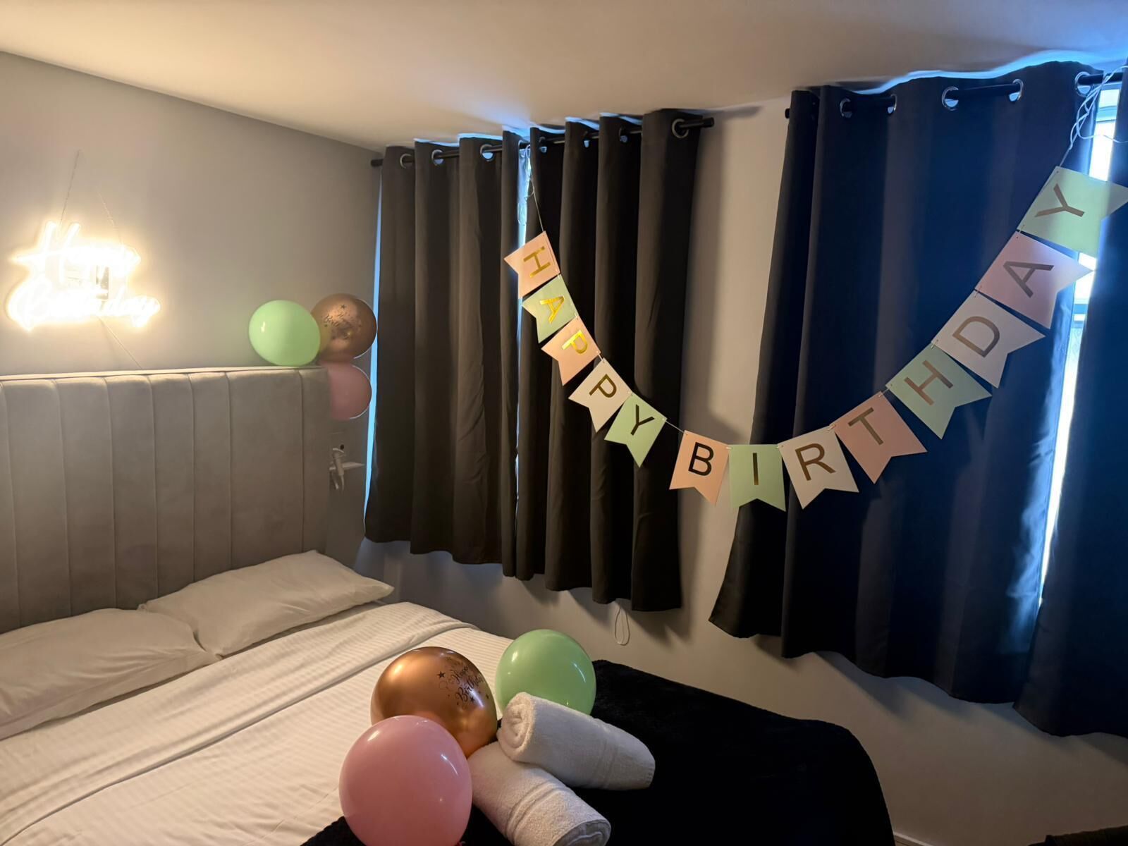 A hotel room with a bed, a wall-mounted light, a curtain, and a birthday banner.