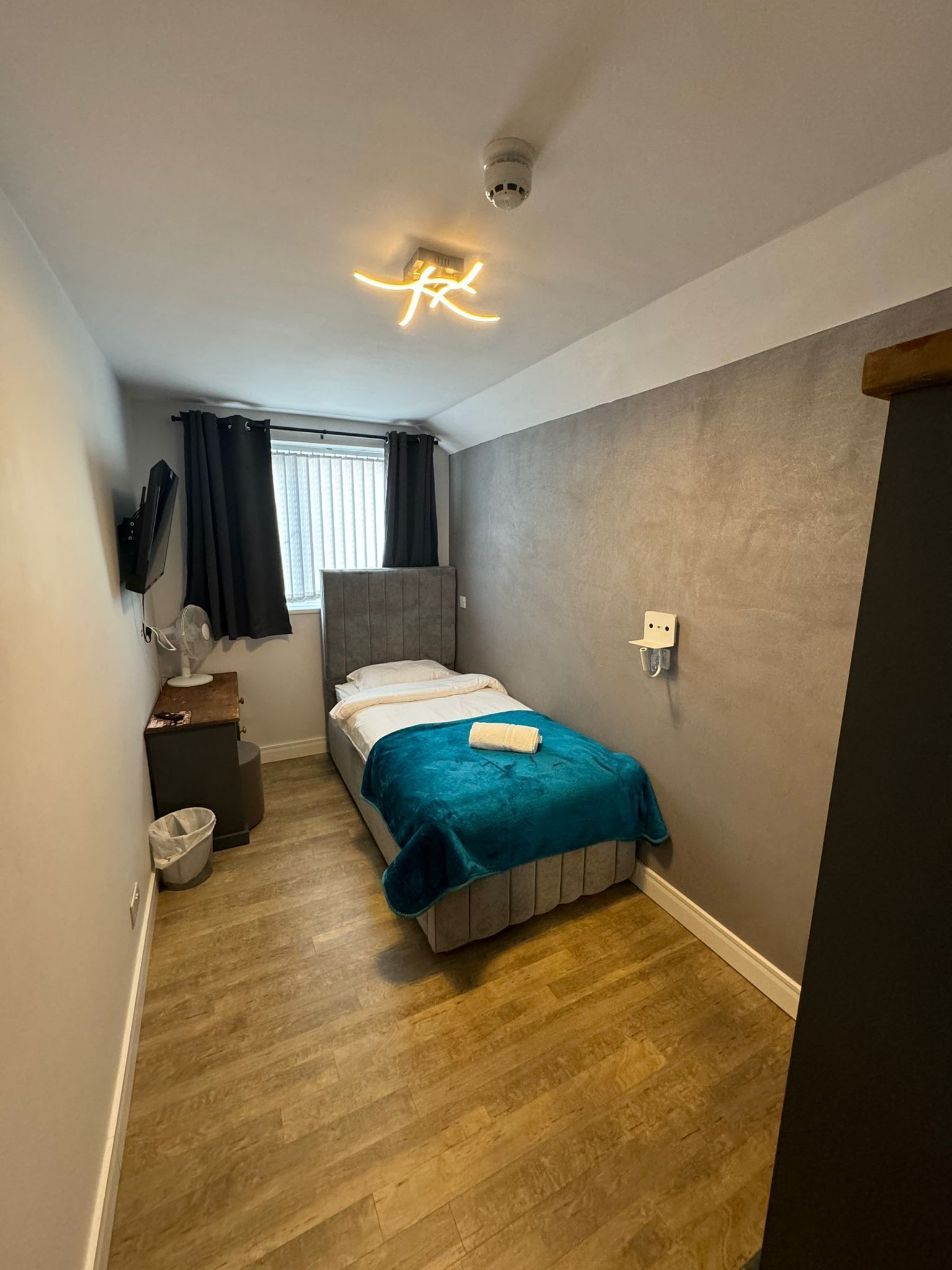 A compact bedroom with a bed, a bedside table, a small desk, a chair, a window with curtains, and a ceiling light fixture.