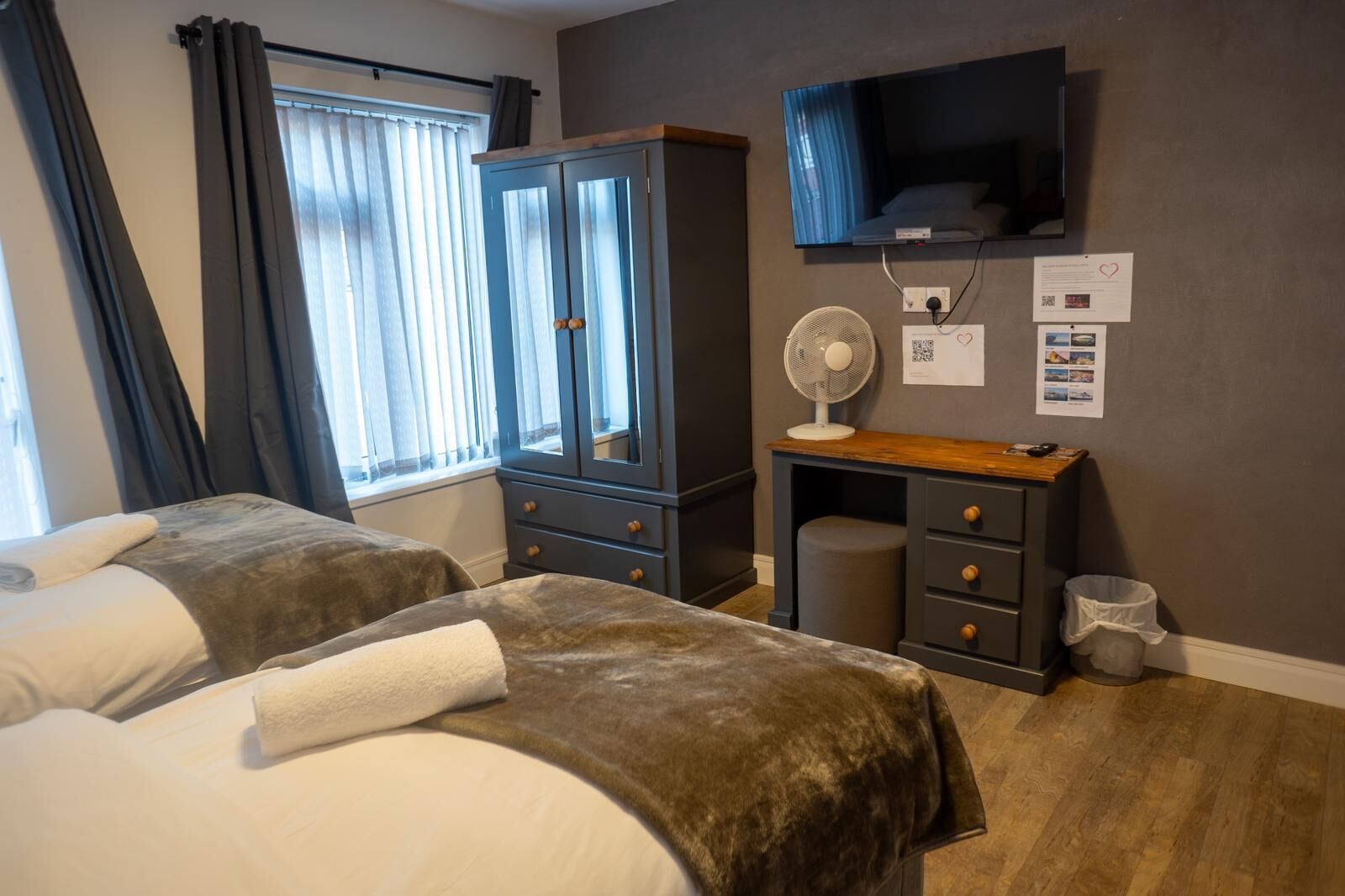A hotel room with two beds, a wardrobe, a desk with a fan, a TV, and a window with curtains.