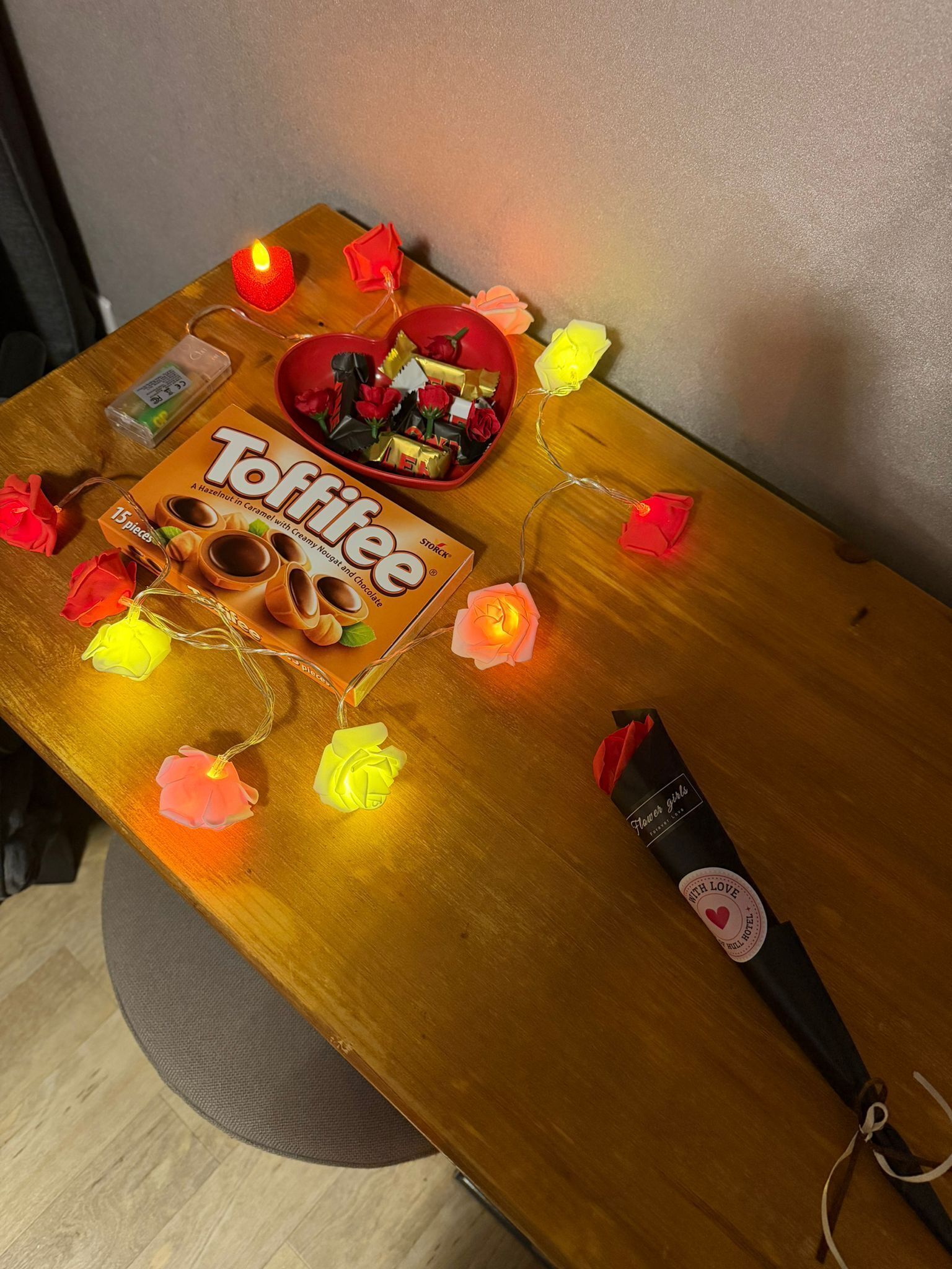 A table with a box of Tofffee, a red heart-shaped bowl with chocolates, and a bottle of wine.