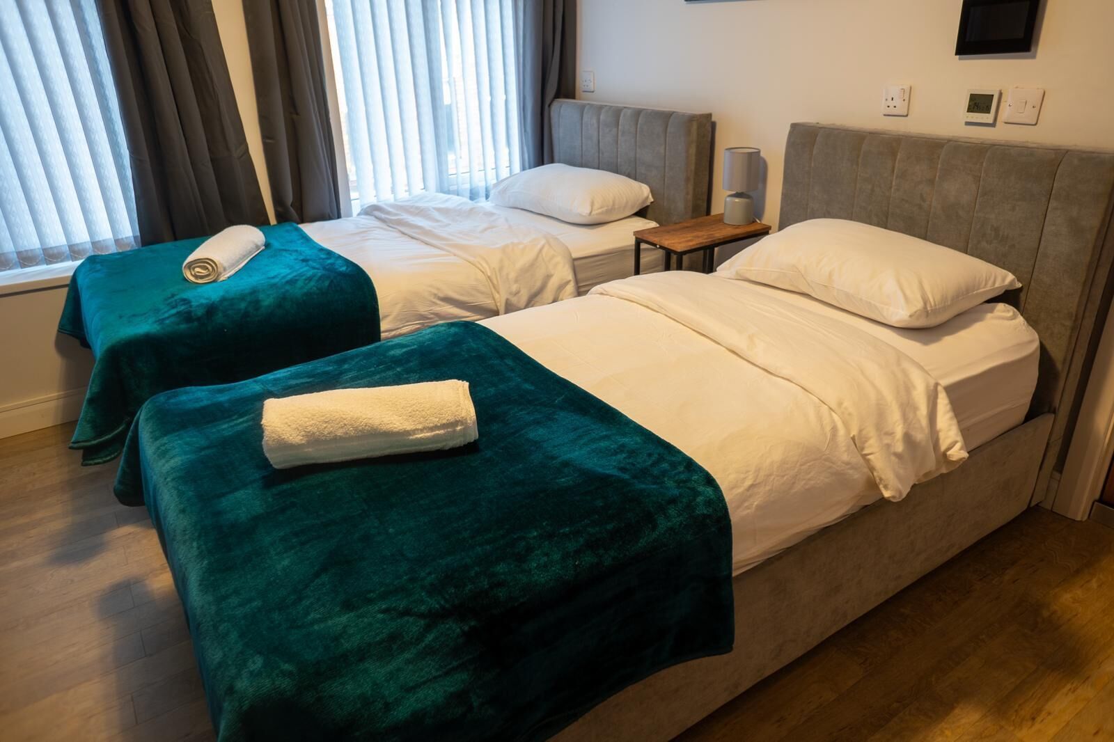 Habitación de hotel con dos camas, cada una con una manta color turquesa y un almohadón blanco. Entre las camas hay una mesita de noche de madera con una lámpara.