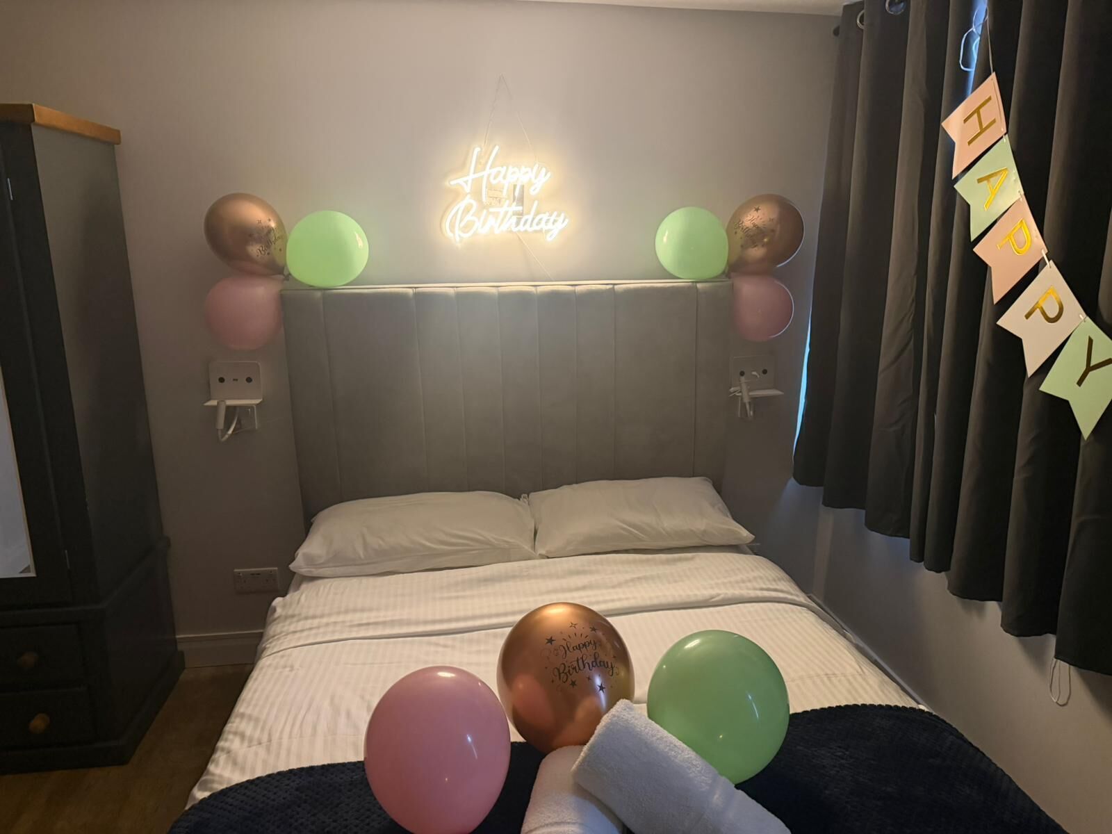 A bedroom with a bed, a neon 'Happy Birthday' sign, balloons, and a 'Happy' banner.