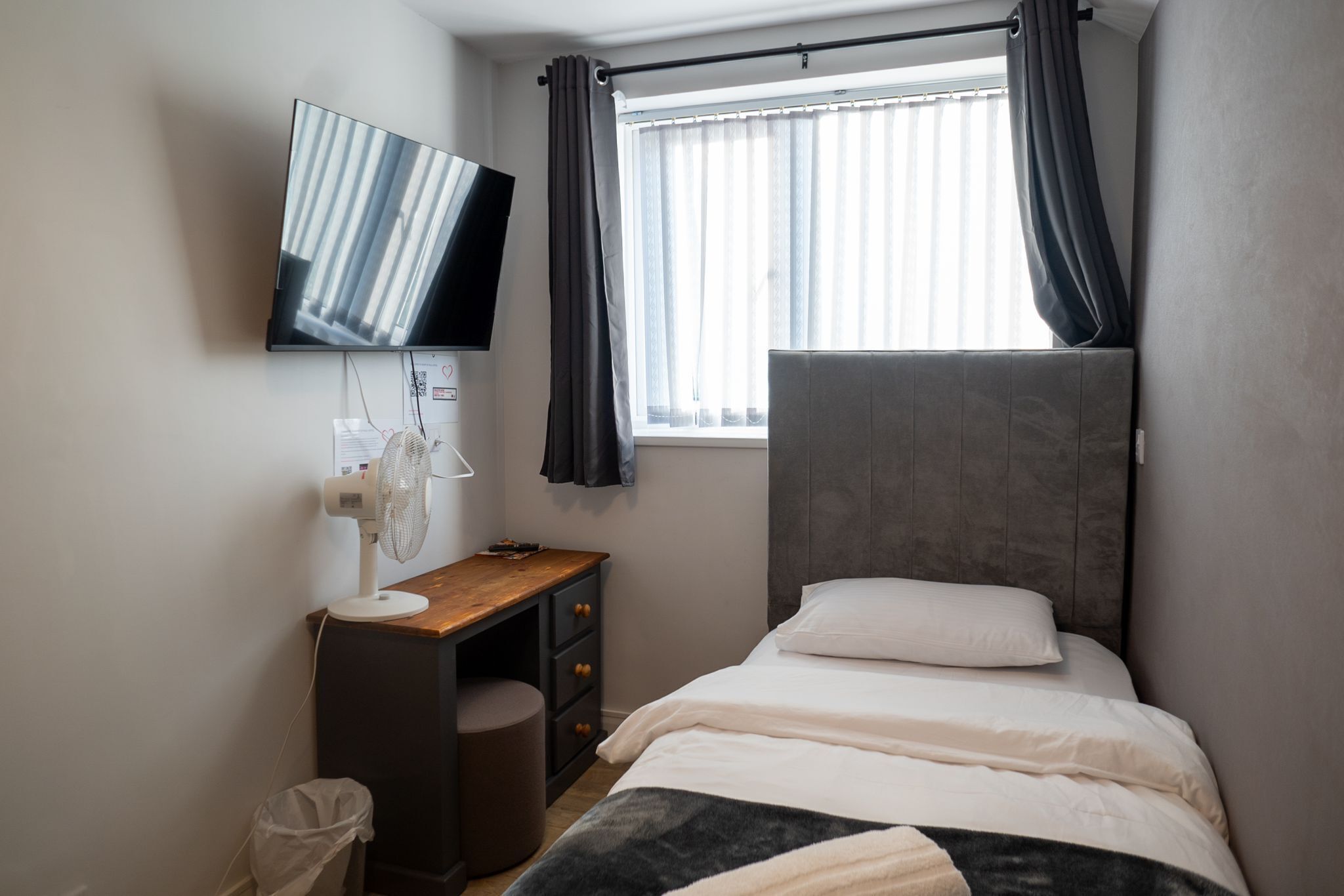 A bedroom with a bed, a desk, a chair, a TV, and a window with curtains.