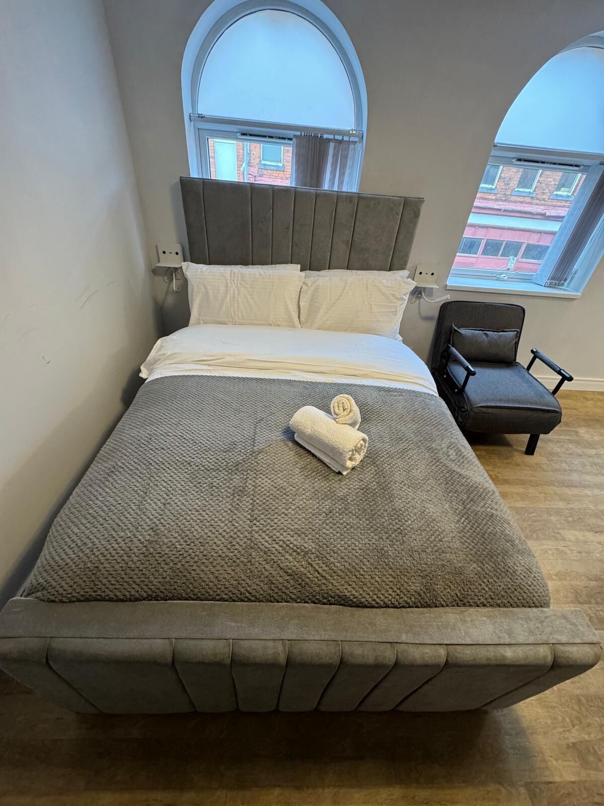 A bed with a headboard, two windows, a chair, and a towel on the bed.