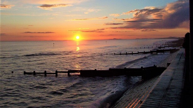 Un coucher de soleil sur une mer calme, avec une jetée en bois qui s’avance dans l’eau.