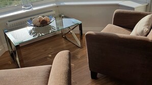 Un salon moderne avec une table basse en verre, deux fauteuils marron et une vue sur l’océan grâce à de grandes fenêtres.
