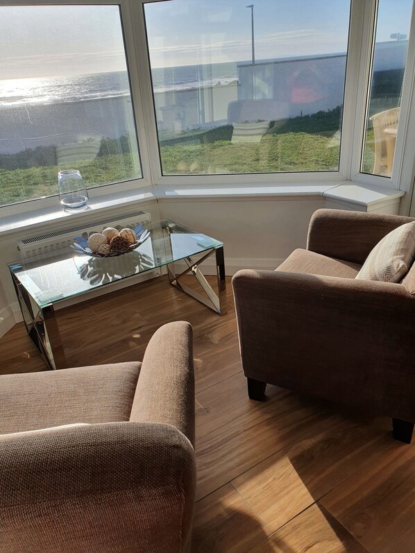 Una sala de estar moderna con una mesa de centro de vidrio, dos sillones marrones y vista al océano a través de amplios ventanales.