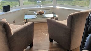 Une pièce avec deux fauteuils en cuir, une table basse en verre sur laquelle se trouve un bol rempli de biscuits, et une vue sur la mer grâce à de grandes fenêtres.