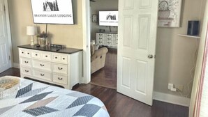 A bedroom with a bed, a dresser, a painting, and a door leading to another room. - Lake Views! Little Bell Bungalow Charmer, Backyard, Game Room, Foosball (Forsyth)