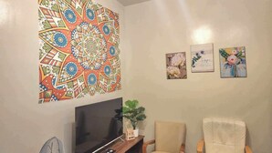 Una sala de estar con un tapiz grande y colorido, un televisor de pantalla plana, dos sillones y tres obras de arte enmarcadas en la pared.