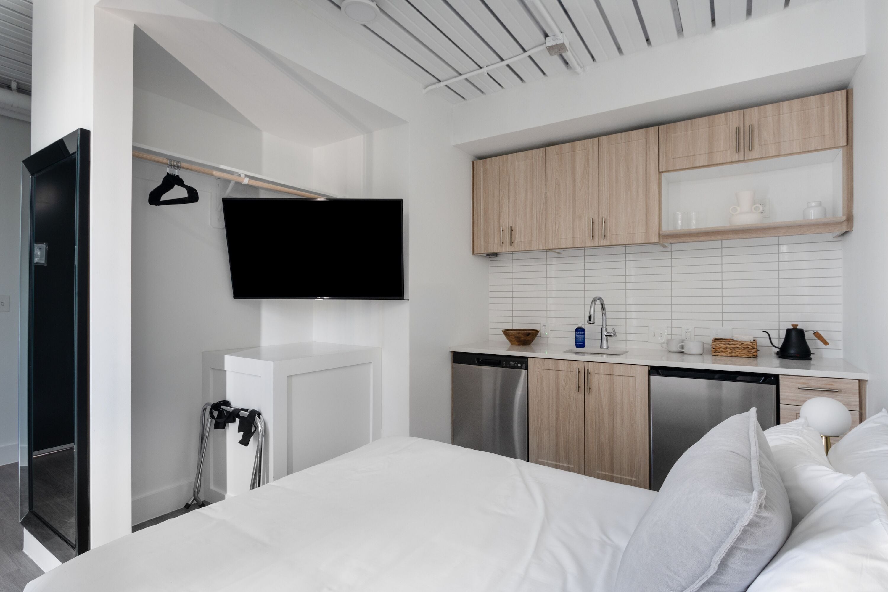 A modern hotel room with a bed, a flat-screen TV mounted on the wall, a kitchen area with wooden cabinets and white tiles, and a small seating area with a table and chairs.